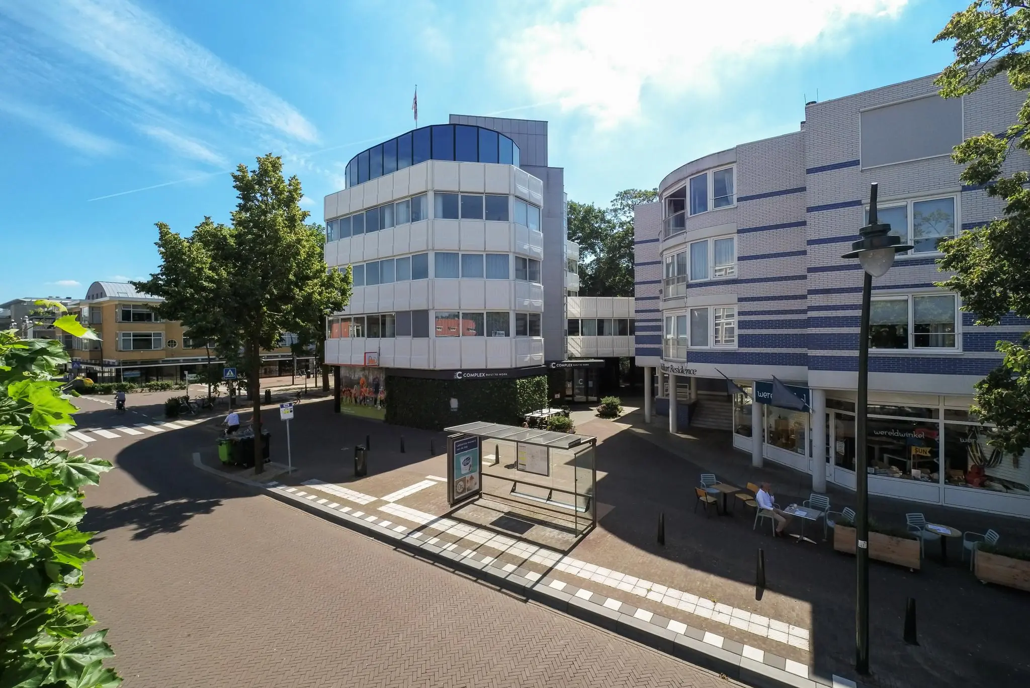 Straatbeeld van de 1e Hogeweg met moderne kantoorgebouwen, winkels, een bushalte en terras onder een blauwe lucht.